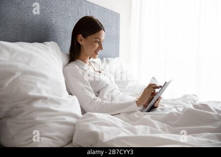 Donna sorridente sdraiata nel letto sotto il piumone, con tablet digitale. Foto Stock