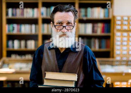 Primo piano ritratto di bello barbuto professore accademico senior o di biblioteca lavoratore, sorridendo e tenendo vecchi libri, mentre in piedi sullo sfondo o Foto Stock