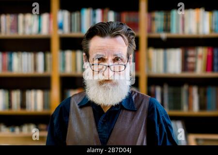 Primo piano ritratto di uomo anziano con barba e occhiali, guardando la macchina fotografica, in piedi su libro negozio sfondo del mercato. Biblioteca, concetto di lettura. Foto Stock