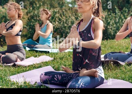 Giovani donne mantenere uno stile di vita sano pratica yoga all'aperto seduti sul tappetino in posa loto mani in preghiera occhi chiusi respirare rilassato vista laterale Foto Stock