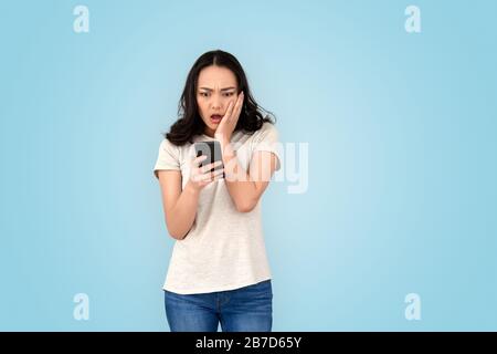 Giovane donna cinese con hairdo in piedi isolato su sfondo grigio che tiene smartphone utilizzando l'applicazione che guarda lo schermo scioccato con messaggio Foto Stock