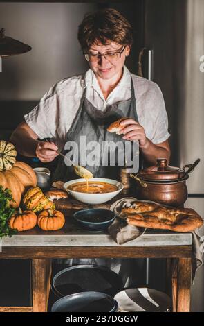 Donna sambuco in grembiule di lino grigio cucina, degustazione e servire autunno stagionale zuppa di crema di zucca in interni cucina moderna. Autunno stagionale comfort foo Foto Stock