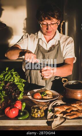 Eldery donna in lino grembiule mettendo il pepe toTurkish tradizionale zuppa di carne Haslama con verdure in cucina moderna con interni. Caduta comfort food, regi Foto Stock