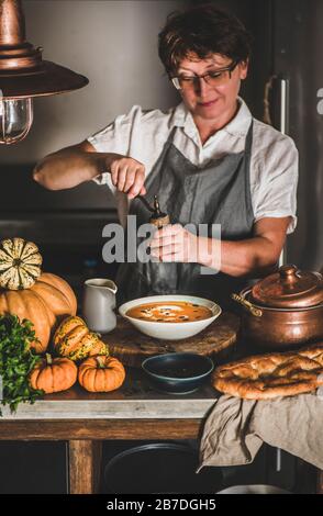 Eldery donna in grigio grembiule biancheria da cucina, degustazione, aggiunta di spezie per stagionale autunno Crema di zucca zuppa in cucina moderna con interni. Caduta comfo stagionali Foto Stock