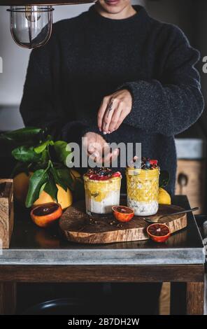 Donna che mette gli ingredienti per Oat Durante La Notte bircher muesli con mango frullato, grano saraceno granola e frutti di bosco freschi in bicchieri su cagna di cemento grigio Foto Stock