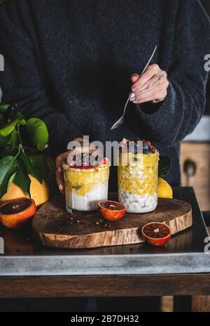 Donna che mangia durante la notte bircher muesli con mango frullato, grano saraceno granola e frutti di bosco freschi in bicchieri su grigio cemento da cucina contatore. Guarisci Foto Stock