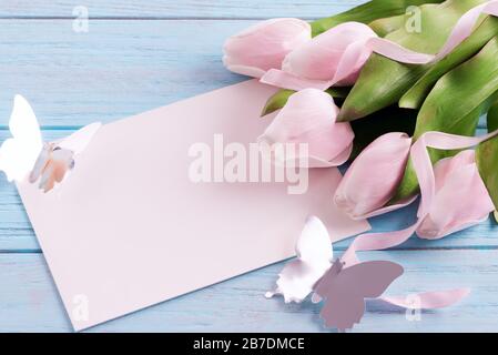 Biglietto di auguri per le vacanze con tulipani rosa tenera, foglio di carta e farfalla artigianale su sfondo di legno blu pastello. Foto Stock