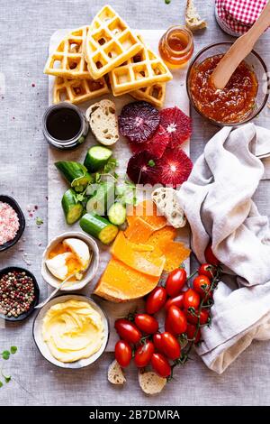 Tavola per la colazione dolce e Salata con baguette, arance sanguigne, burro, formaggio, caffè, cetrioli, gouda, miele, marmellata, sciroppo d'acero, arancia, pepe, rosa s. Foto Stock