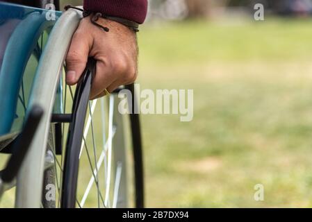 Mano di un uomo adulto paralizzato su una sedia a rotelle che indossa una felpa di colore rosso scuro in un parco Foto Stock
