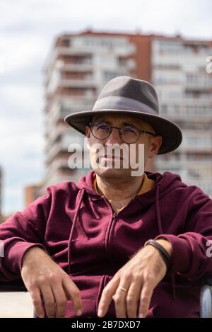 Uomo caucasico con occhiali e cappello depressivo e triste in una sedia a rotelle solitaria in un parco con il cielo e alcuni edifici sullo sfondo con lo spazio Foto Stock