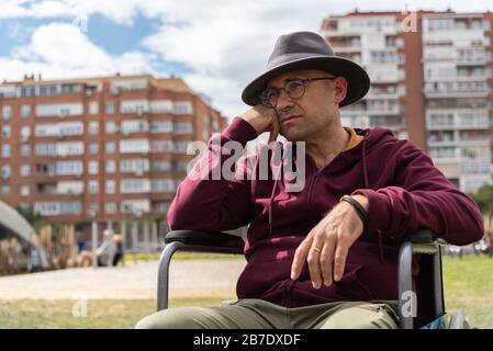 Uomo caucasico con occhiali e cappello depressivo e triste in una sedia a rotelle solitaria in un parco con il cielo e alcuni edifici sullo sfondo con lo spazio Foto Stock