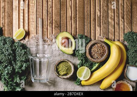 Vetro per frullati vegani verdi sani, paglia di vetro. Ingredienti sopra. Kale, banane, avocado, calce, latte non-diario, miele, polvere di matcha e semi o Foto Stock