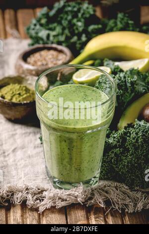 Bicchiere di frullato vegano verde sano, paglia di vetro. Ingredienti sopra. Kale, banane, avocado, calce, latte non-diario, polvere di matcha e semi sul legno Foto Stock