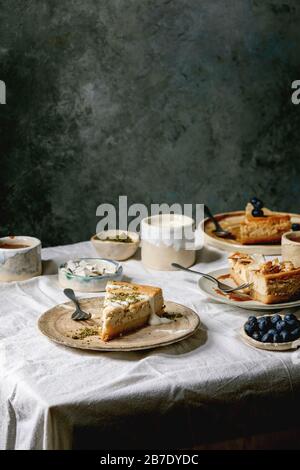 Pezzi di cheesecake con condimenti diversi su piatti di ceramica e varietà di salse in ciotole di ceramica su tovaglia bianca. Dessert per colazione Foto Stock