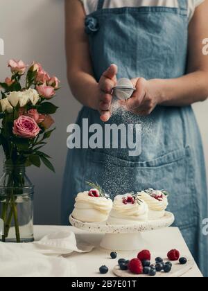 Donna in un grembiule spolga lo zucchero a velo attraverso un setaccio su mini Pavlova torte con frutti di bosco freschi. Composizione verticale con piano in marmo bianco an Foto Stock