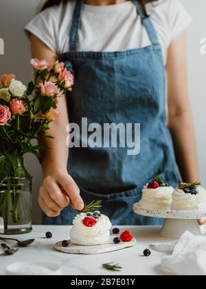 Donna in un grembiule tiene rosmarino con le dita per decorare mini Pavlova torta con frutti di bosco freschi. Composizione verticale con piano in marmo bianco an Foto Stock