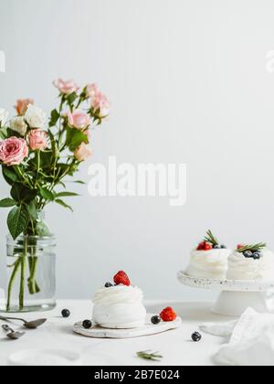 Mini torte Pavlova con frutti di bosco freschi e rosmarino su tavolo in marmo bianco con fiori in bottiglia di vetro e parete bianca sullo sfondo. COM. Verticale Foto Stock