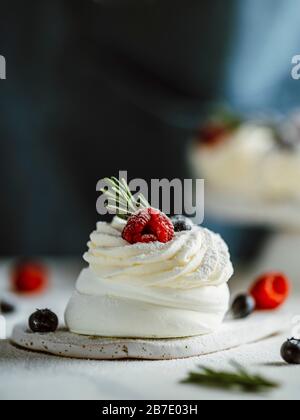 Mini torta Pavlova con frutti di bosco freschi e rosmarino verde fresco. Primo piano a composizione verticale con piano in marmo bianco e sfondo scuro di grembiule. Foto Stock