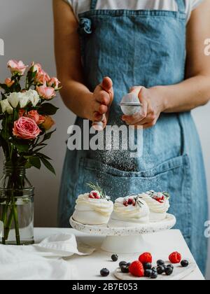 Donna in un grembiule spolga lo zucchero a velo attraverso un setaccio su mini Pavlova torte con frutti di bosco freschi. Composizione verticale con piano in marmo bianco an Foto Stock