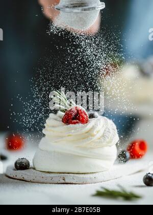 Donna spruzza lo zucchero a velo attraverso un setaccio su mini Pavlova torta decorata frutti di bosco freschi e rosmarino. Composizione verticale. Foto Stock