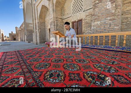Uomo uzbeko seduto sul tappeto e leggendo nel cortile del complesso religioso Chor Bakr, a Bukhara, Uzbekistan Foto Stock