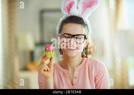 casalinga moderna sorridente in una blusa rosa e spighe conigliate di pasqua con i vetri che tengono l'uovo rosso di pasqua nella tazza verde dell'uovo alla casa moderna in da di primavera soleggiato Foto Stock