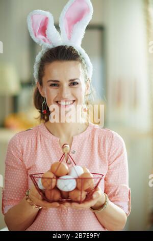 happy elegante casalinga di mezza età in una blusa rosa e spighe conigliate di pasqua tenendo cesto con uova a casa moderna in sole primavera giorno. Foto Stock