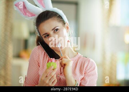 giovane donna di 40 anni in una blusa rosa e spighe conigliate di pasqua che parlano su uno smartphone mentre mangiano l'uovo di pasqua rosso in una tazza di uovo verde con cucchiaio in th Foto Stock