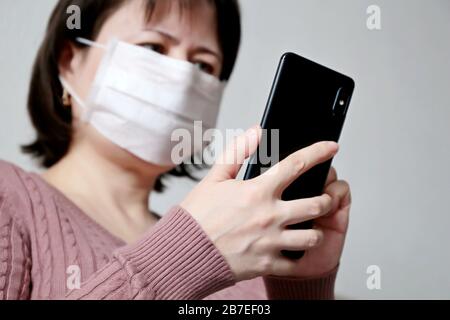 Donna in maschera medica con smartphone, telefono cellulare in primo piano in mani femminili. Concetto di malattia, febbre, raffreddore e influenza, ricerca del sintomo di coronavirus Foto Stock