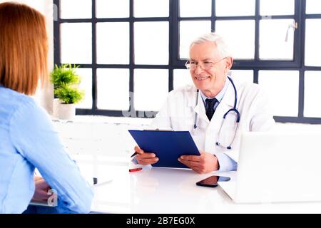 Colpo di medico di mezza età che parla con il paziente mentre si siede alla scrivania in camera del medico. Foto Stock