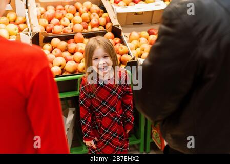 Una bambina di circa 5 ha gettato un tantrum in un supermercato davanti ai suoi genitori. Il bambino urla e grida, mendicando dolci da mamma e papà sullo sfondo di frutta e mele. Foto Stock