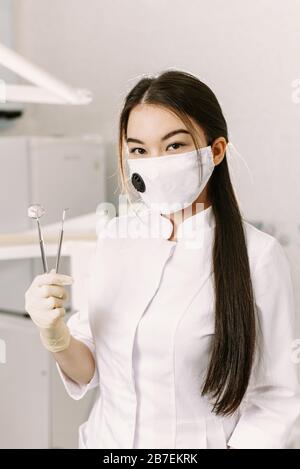 Allegra dentista femmina sorridente al suo ufficio. Bella studentessa donna in una maschera protettiva in odontoiatria si trova nell'ufficio dentale e tiene strumenti professionali. Foto Stock