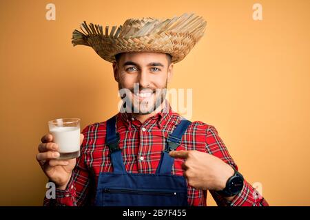 Giovane contadino uomo che indossa cappello di campagna bere latte biologico dalla fattoria con la faccia sorpresa che punta il dito a se stesso Foto Stock