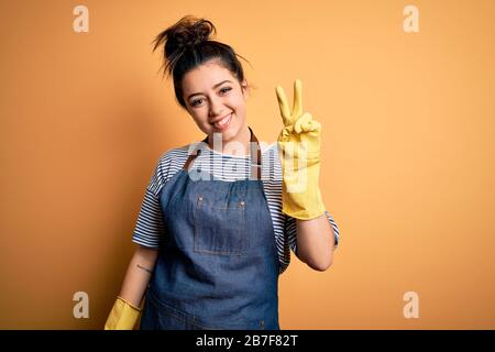 Giovane donna brunette pulitore indossare guanti di pulizia su sfondo giallo sorridente con faccia felice che si annida alla fotocamera facendo segno di vittoria. Numero Foto Stock