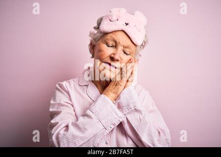 Donna anziana bella che indossa la maschera del sonno e pajama su sfondo rosa isolato dormire stanco sognare e posare con le mani insieme mentre smilin Foto Stock