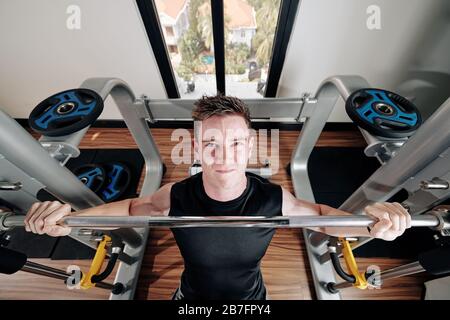 Bel giovane uomo forte che guarda in alto quando si siede su panchina sollevando pesante barbell, vista dall'alto Foto Stock