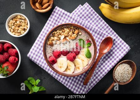 Ciotola per frullati rosa con lampone banana mandorle di chia semi e granola su sfondo nero. Cibo sano e pulito. Superfood di frutta liscio Foto Stock
