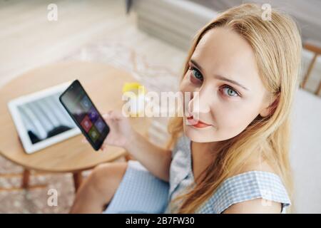 Bella giovane donna bionda con occhi verdi guardando la fotocamera quando si utilizza l'applicazione smart home sul suo telefono Foto Stock