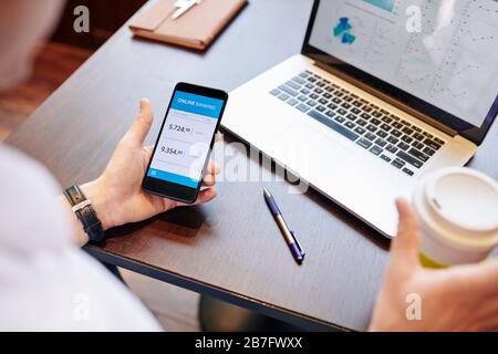 Uomo d'affari che beve caffè e controlla i tassi di cambio tramite l'applicazione bancaria per smartphone quando lavora con il report finanziario Foto Stock