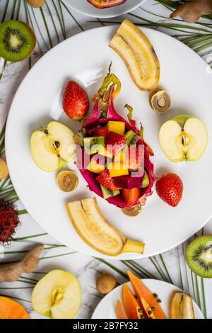 Mix di diversi frutti tropicali freschi maturi su piatto bianco e sul tavolo con foglie di palma verde. Colazione esotica, snack deliziosi. Estate, vacanza e Foto Stock