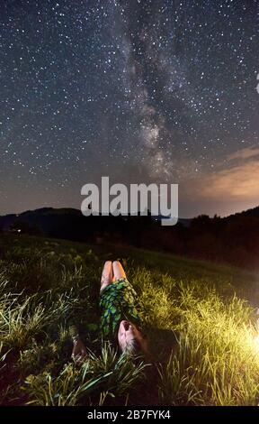 Fotografia verticale di una ragazza che giace in erba estiva di notte osservando il bel cielo pieno di stelle e Via Lattea in montagna. Concetto di unità con la natura Foto Stock