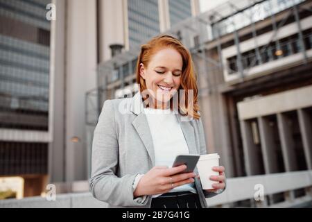 Giovane donna di successo che tiene una tazza da caffè da asporto e utilizza lo smartphone mentre si trova all'aperto Foto Stock