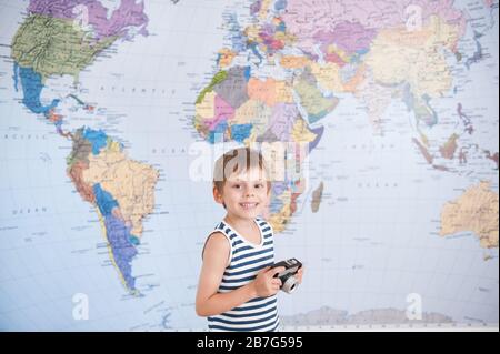 felice piccolo delizioso bambino in canotta marinaio a righe che tiene la macchina fotografica del film d'epoca sullo sfondo della mappa del mondo Foto Stock