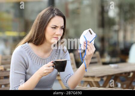 Una giovane donna dubbia che guarda sospettosa alla maschera facciale protettiva sulla terrazza di un bar Foto Stock