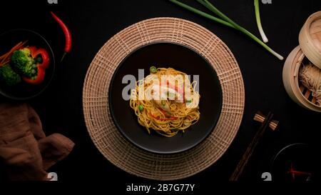 Vista dall'alto di Schezwan Noodles o Chow Mein con verdure, pollo e salsa di peperoncino serviti in ciotola nera e ingredienti su tavolo nero Foto Stock