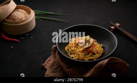 Espresso di Schezwan Noodles o Chow Mein con verdure e pollo serviti in una ciotola nera e ingredienti sul tavolo nero Foto Stock