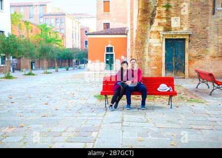 Coppia turistica multirazziale, asiatica caucasica, all'inizio degli anni Cinquanta seduta insieme su panchina rossa in piazza cittadina vuota a Venezia, Italia Foto Stock