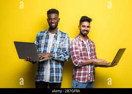 Ritratto di due giovani felici che tengono il computer portatile mentre si sta in piedi e celebrano isolato su sfondo giallo Foto Stock