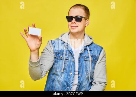 Elegante uomo caucasico in jeans e occhiali da sole su sfondo giallo. Contiene una carta bianca. Spazio di copia. Primo piano. Foto Stock