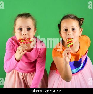 Felicità e concetto di dessert. Sorelle con lollipopps rotondi e lunghi. Le ragazze con i volti affamati posano con le caramelle su sfondo verde. I bambini mangiano grandi caramelle dolci colorate. Foto Stock